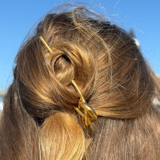 A close-up back view of a person's hair styled in a half-up bun with a gold twisted hair claw clip holding it in place, set against a clear blue sky.