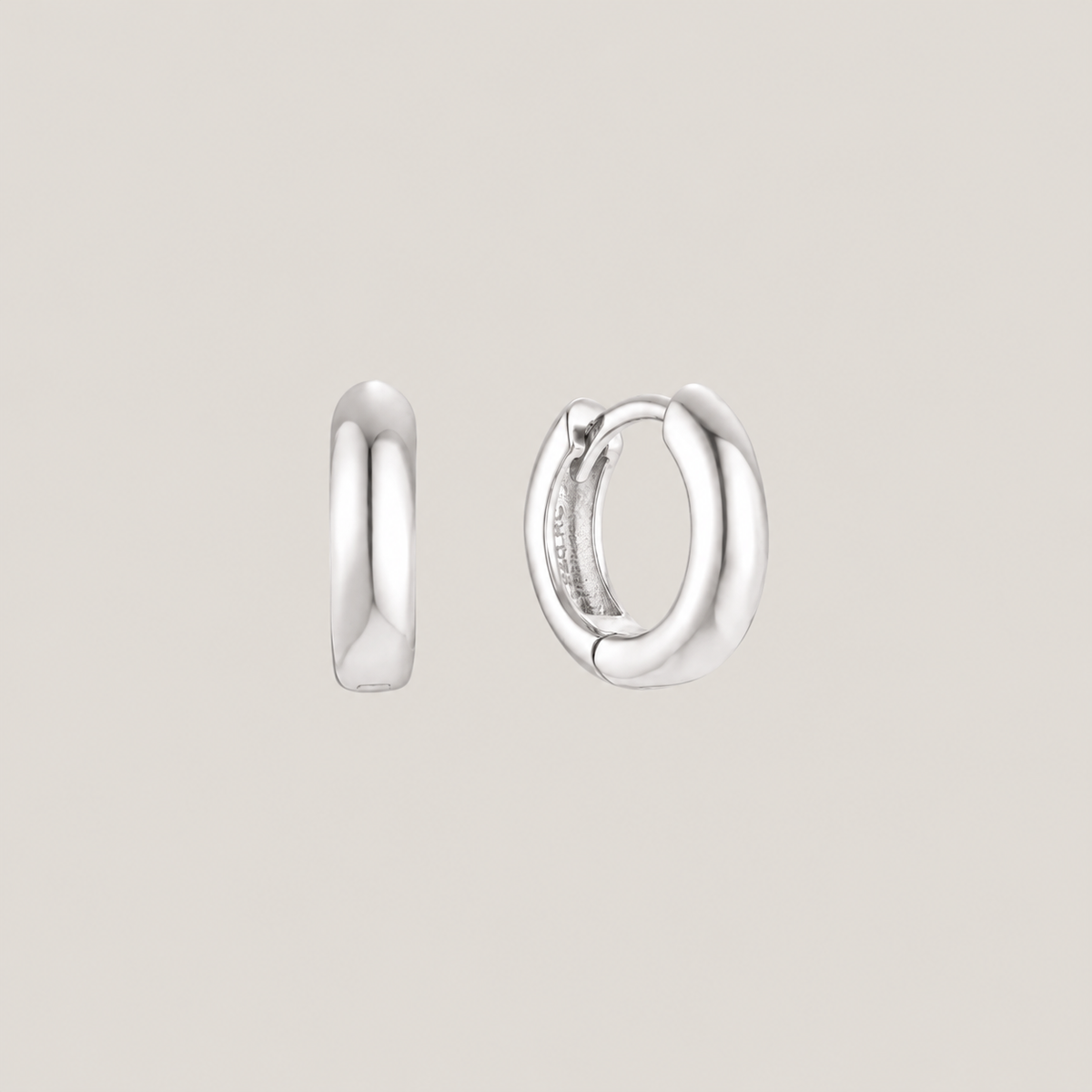 A pair of polished silver huggie hoop earrings shown from two angles against a solid light beige background.