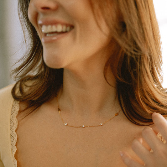 A close-up lifestyle shot of a woman wearing a delicate gold necklace with small white bead accents and a cream lace-trimmed top.
