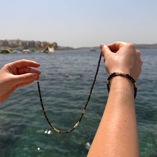 A person outdoors by the water holds up a beaded necklace made of dark brown and gold beads. The person's hands and forearms are visible against a background of blue sea and sky.