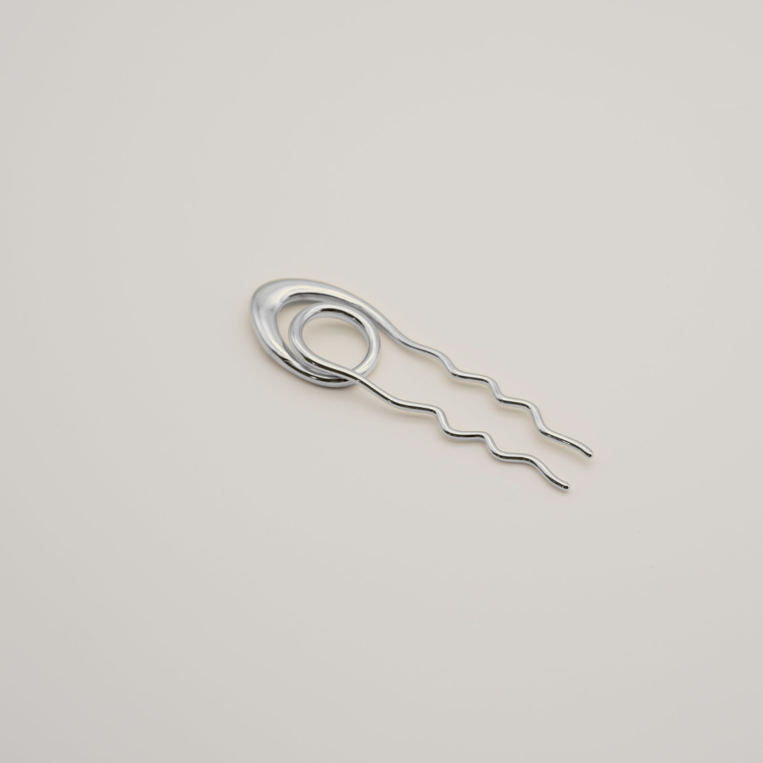 A silver metal hair pin with a coiled top and three wavy straight prongs, shown on a plain light beige background.