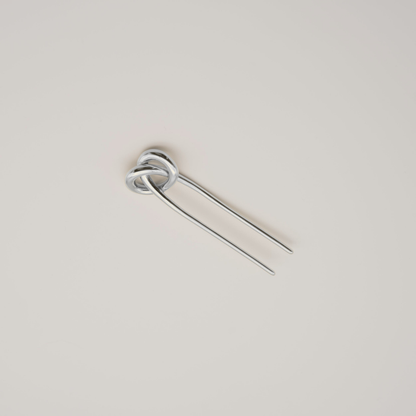 A silver metal hair pin with a knotted loop at the top and two long straight prongs, shown on a plain light beige background.