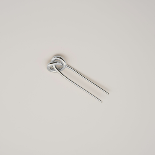A silver metal hair pin with a knotted loop at the top and two long straight prongs, shown on a plain light beige background.