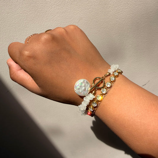 A close-up of a person's wrist and hand wearing a stack of bracelets against a neutral light gray wall. The bracelets include a gold toggle clasp with a large white iridescent pearl, a beaded bracelet with white stones, and a gold beaded bracelet with clear rhinestones 