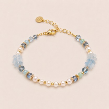 A delicate gold chain bracelet featuring white freshwater pearls, faceted light blue and aquamarine beads, and small gold spacer beads, displayed on a light beige background.