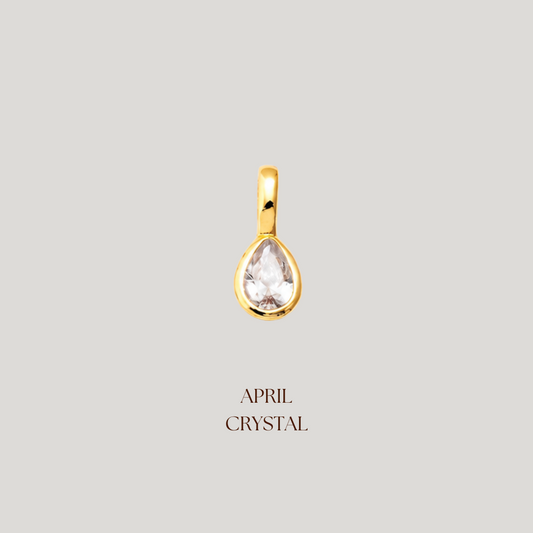 Gold teardrop-shaped pendant with a clear gemstone on a light gray background, labeled 'April Crystal'.