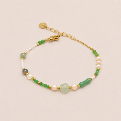 A delicate gold chain bracelet featuring white pearls, green beads, and a large light green stone centerpiece, displayed on a soft beige background.