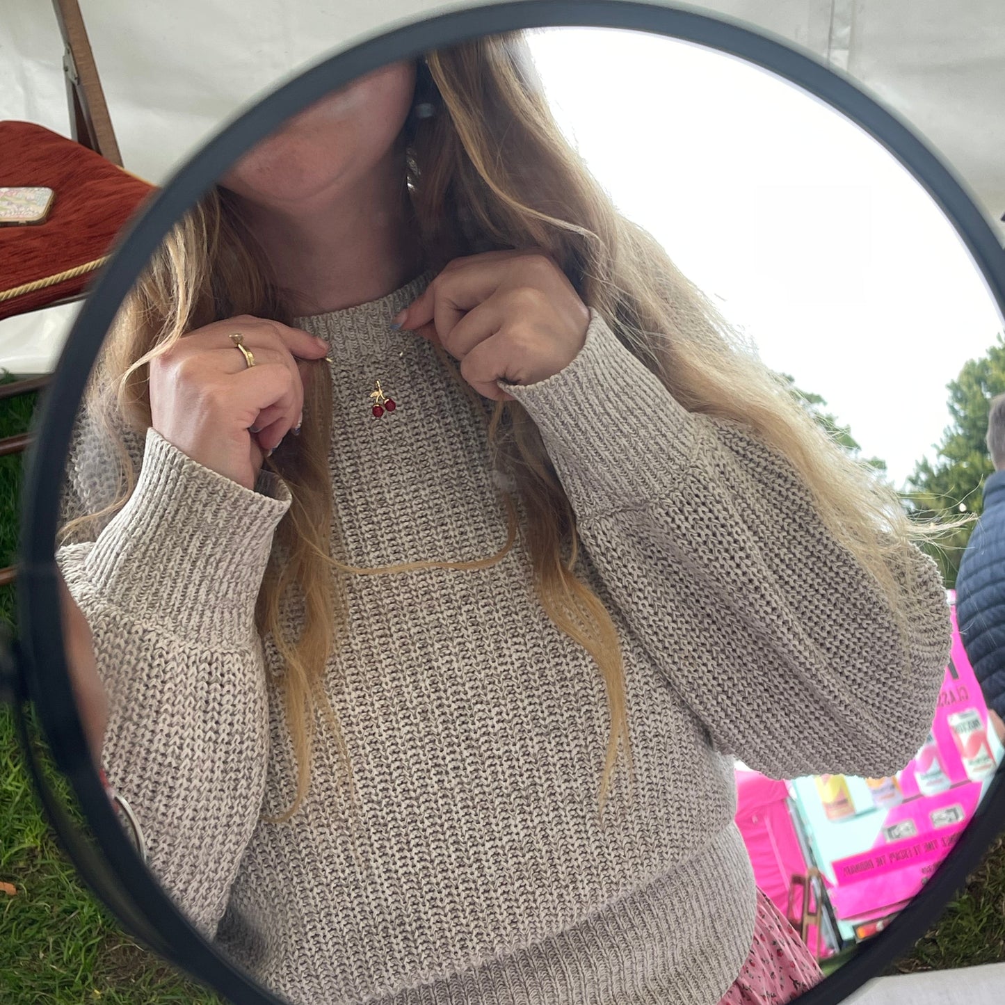 Person wearing a beige knit sweater looking into a round mirror wearing a cherry charm.