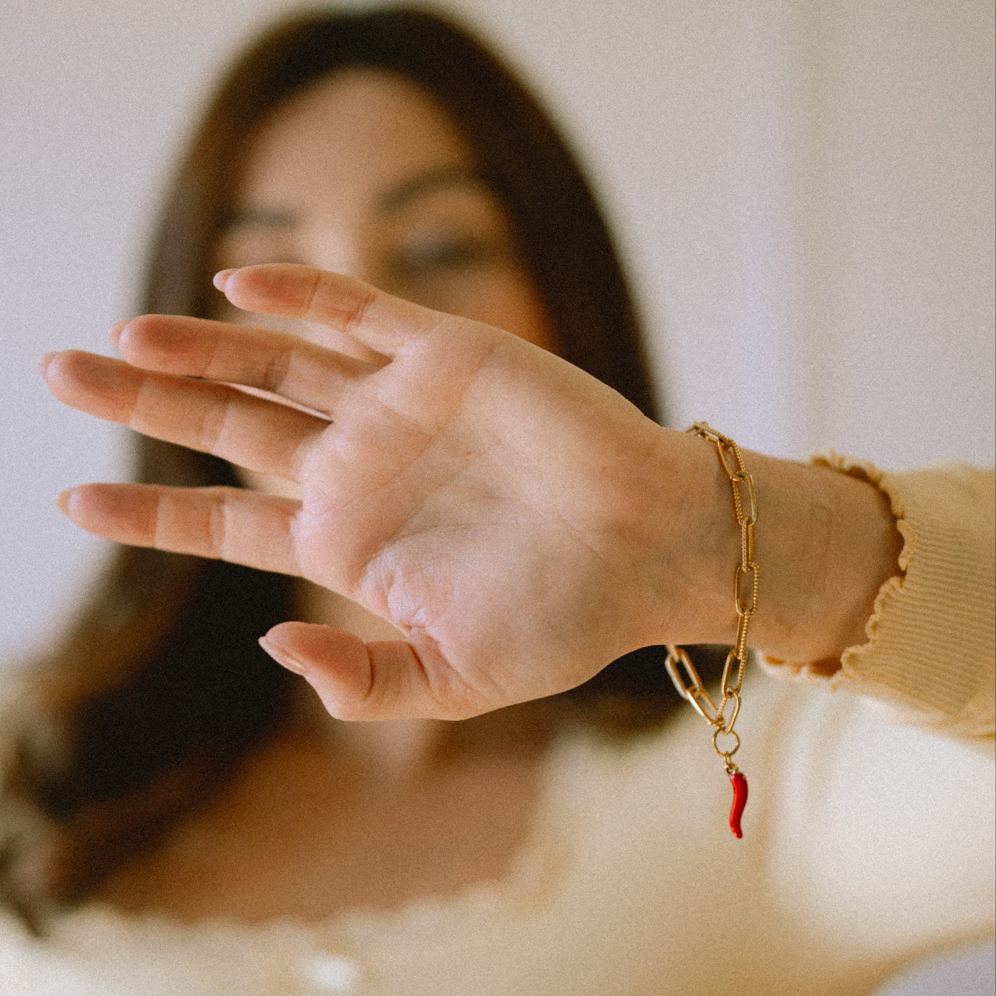 Woman wearing a gold bracelet with a red charm, holding her hand out in front of her face.