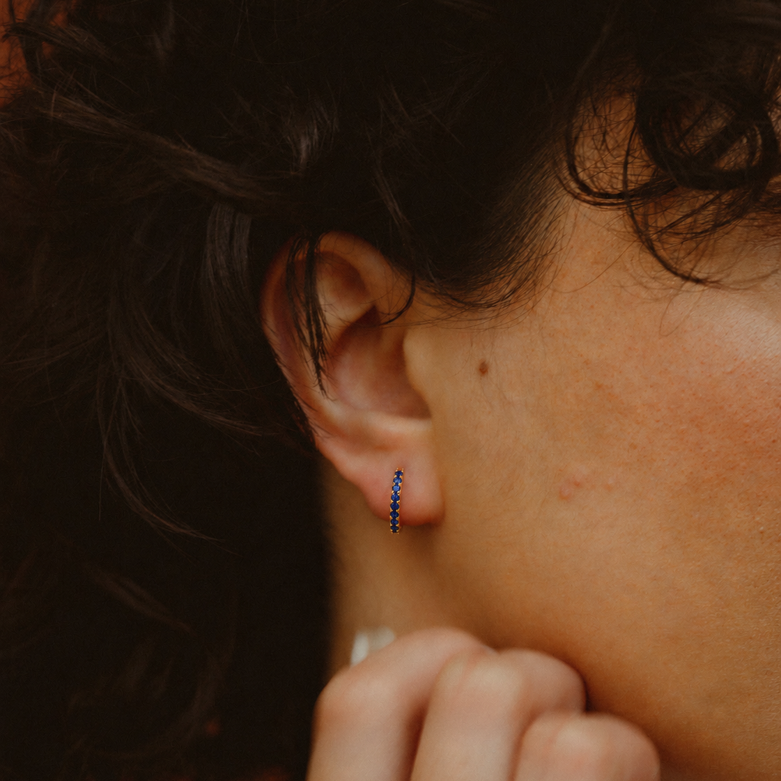 A close-up of a person's ear wearing a small vertical huggie hoop earring with tiny dark blue beads.