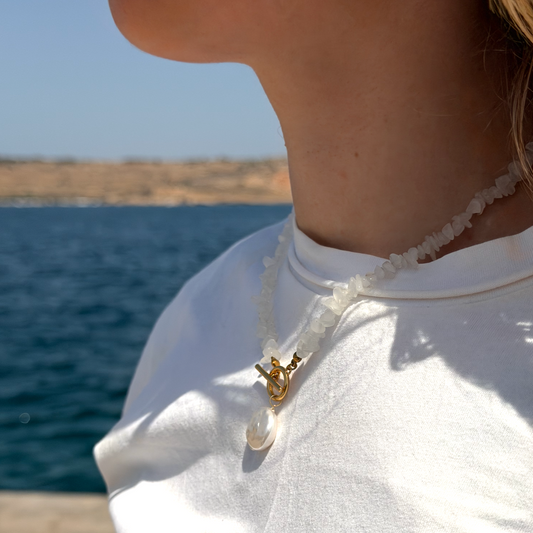 A close-up lifestyle shot of a person outdoors by the water wearing a white beaded necklace with a gold anchor charm and a white pearl pendant.