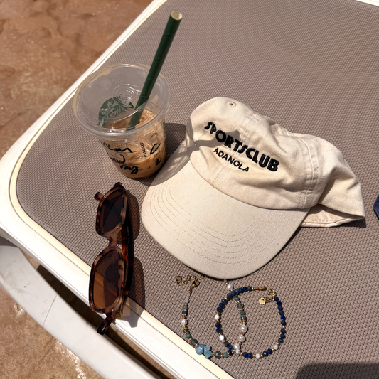 A beige baseball cap with 'SPORT CLUB ADANOLA' printed in black on the front, styled on a gray mesh tray alongside a Starbucks iced coffee, tortoiseshell sunglasses, and beaded bracelets.