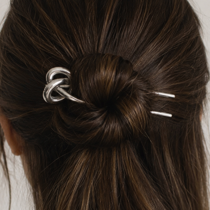 A close-up of a dark brown hair bun secured with a silver metallic knot hair claw clip, with two small silver bar pins holding the sides, shown on a model.